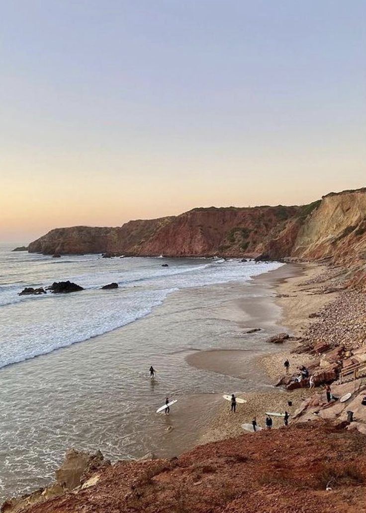 Des surfeurs traversant une large baie au crépuscule sous des falaises rouges, Algarve